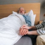 Close-up of unrecognizable loving young woman child holding hand of sick aged father lying in hospital bed.